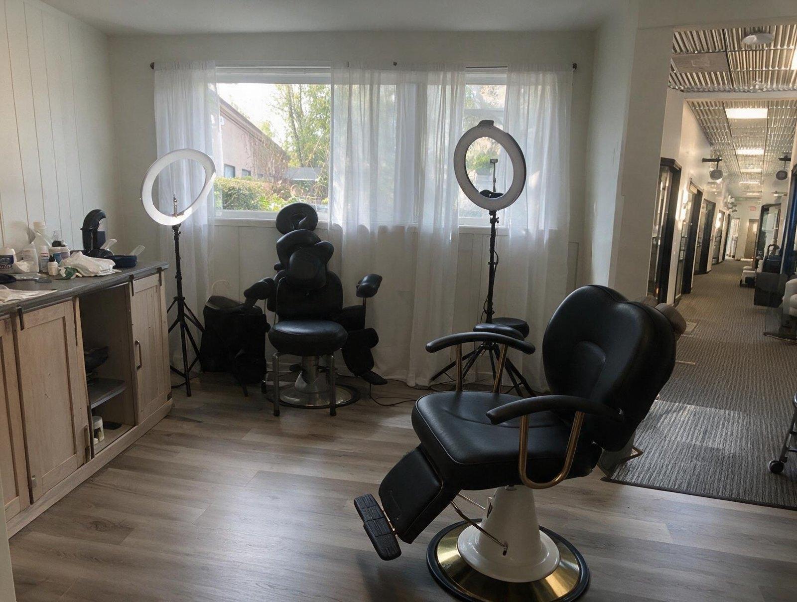 Treatment room with black reclining chairs, two ring lights, a wooden cabinet of supplies and sheer curtained windows