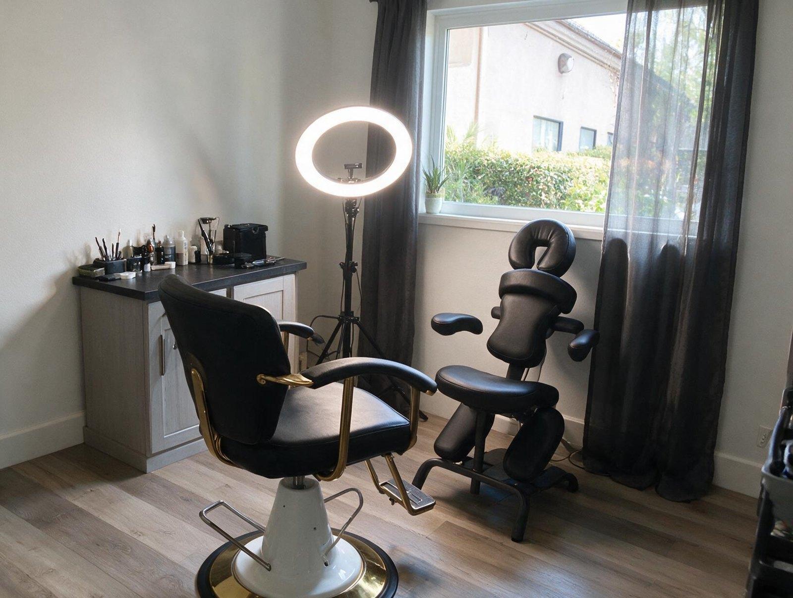 Treatment room with a black and gold swivel chair, a ring light, a black massage chair and a supply table by a window.
