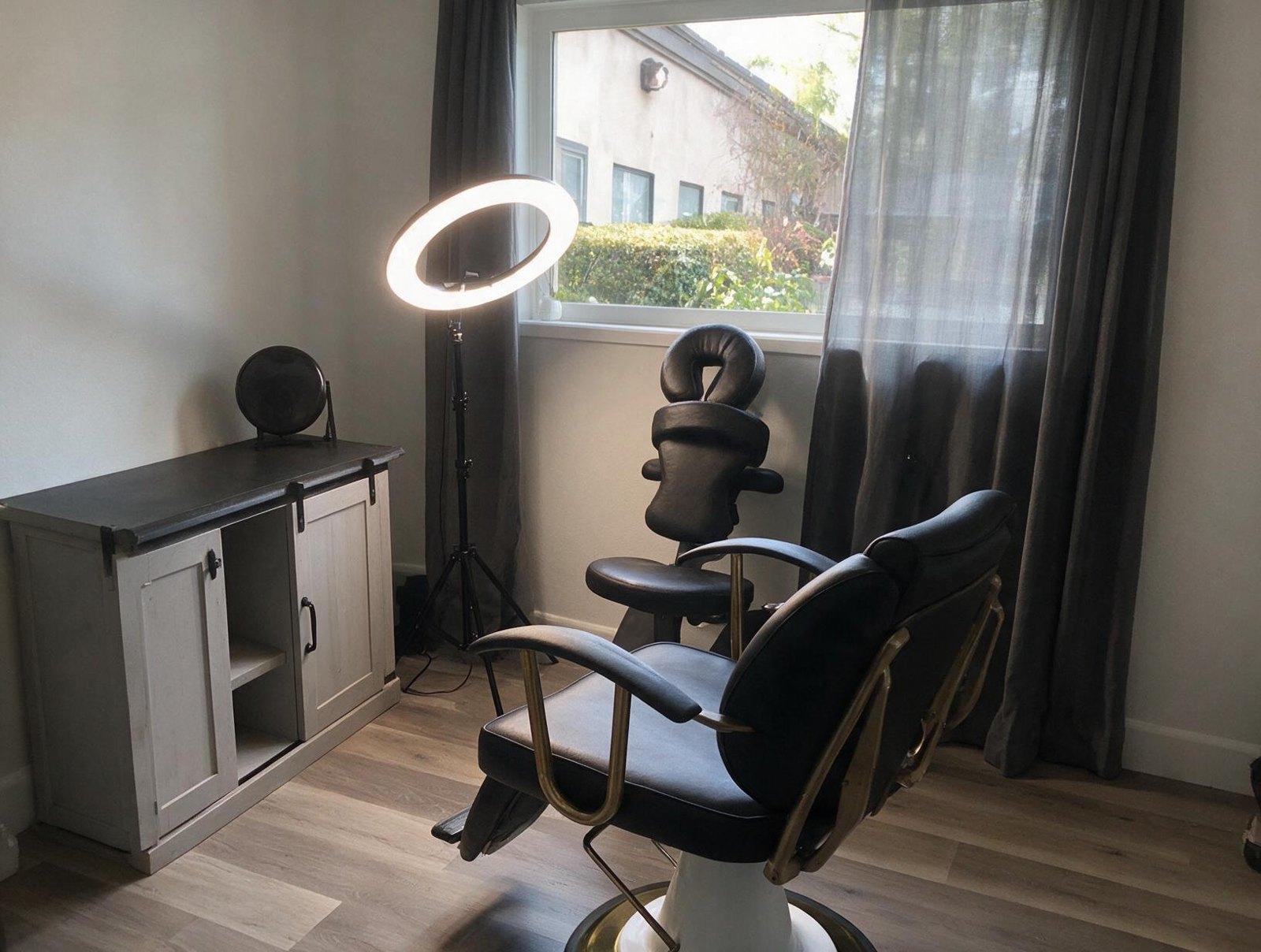 Empty treatment room with a black leather chair, ring light, face-rest chair, grey curtains, a white cabinet and a window.
