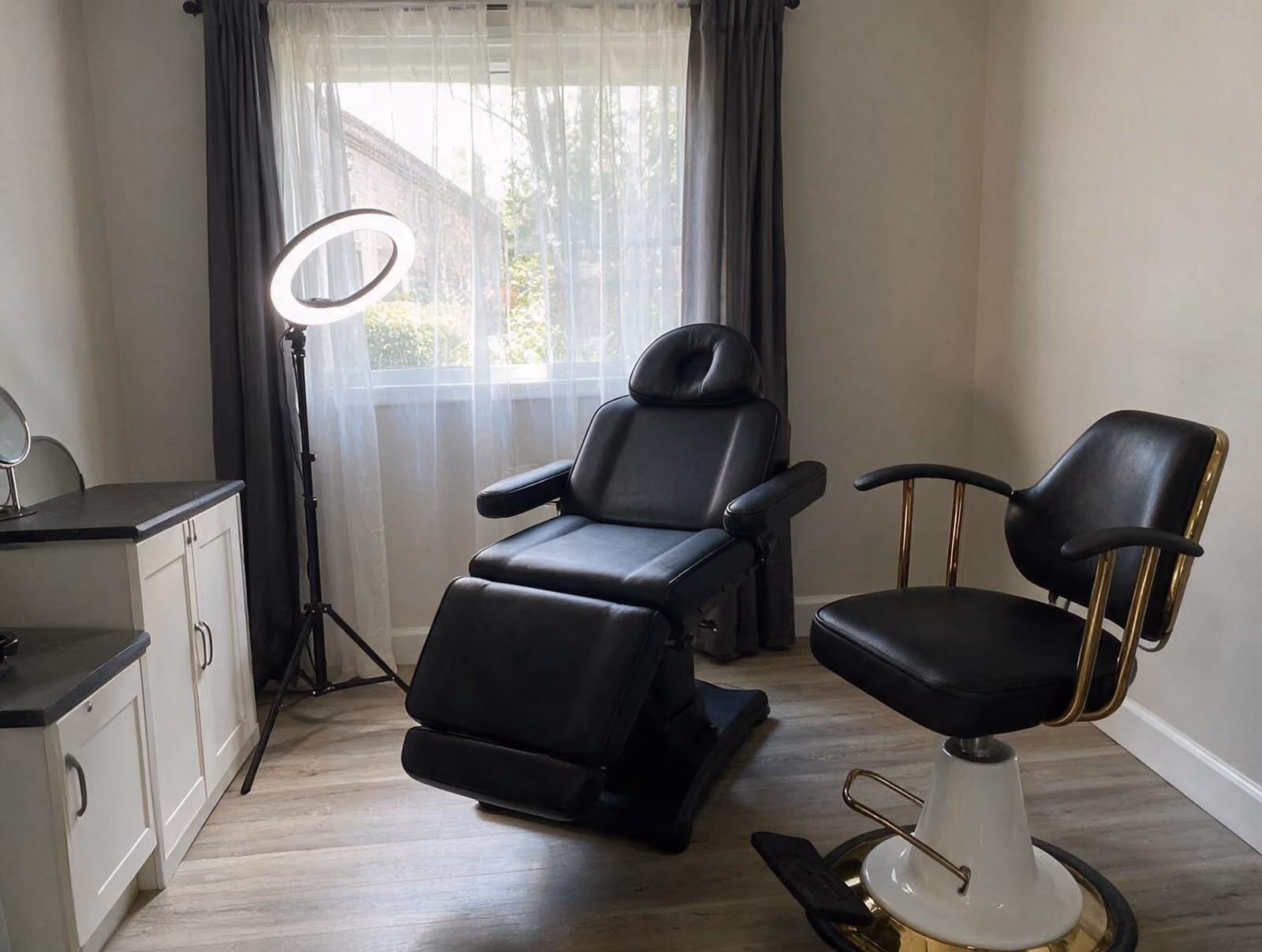 Empty treatment room with a black reclining chair, a gold-trimmed swivel chair and a ring light by a sheer-curtained window