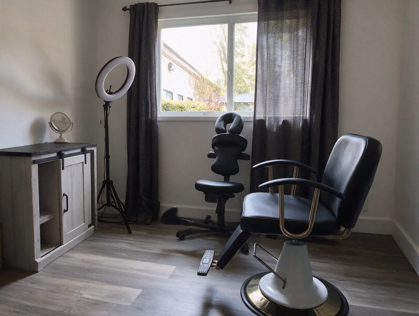 Quiet treatment room with a black salon chair, a face-cradle massage chair, a ring light and a small mirror by a window