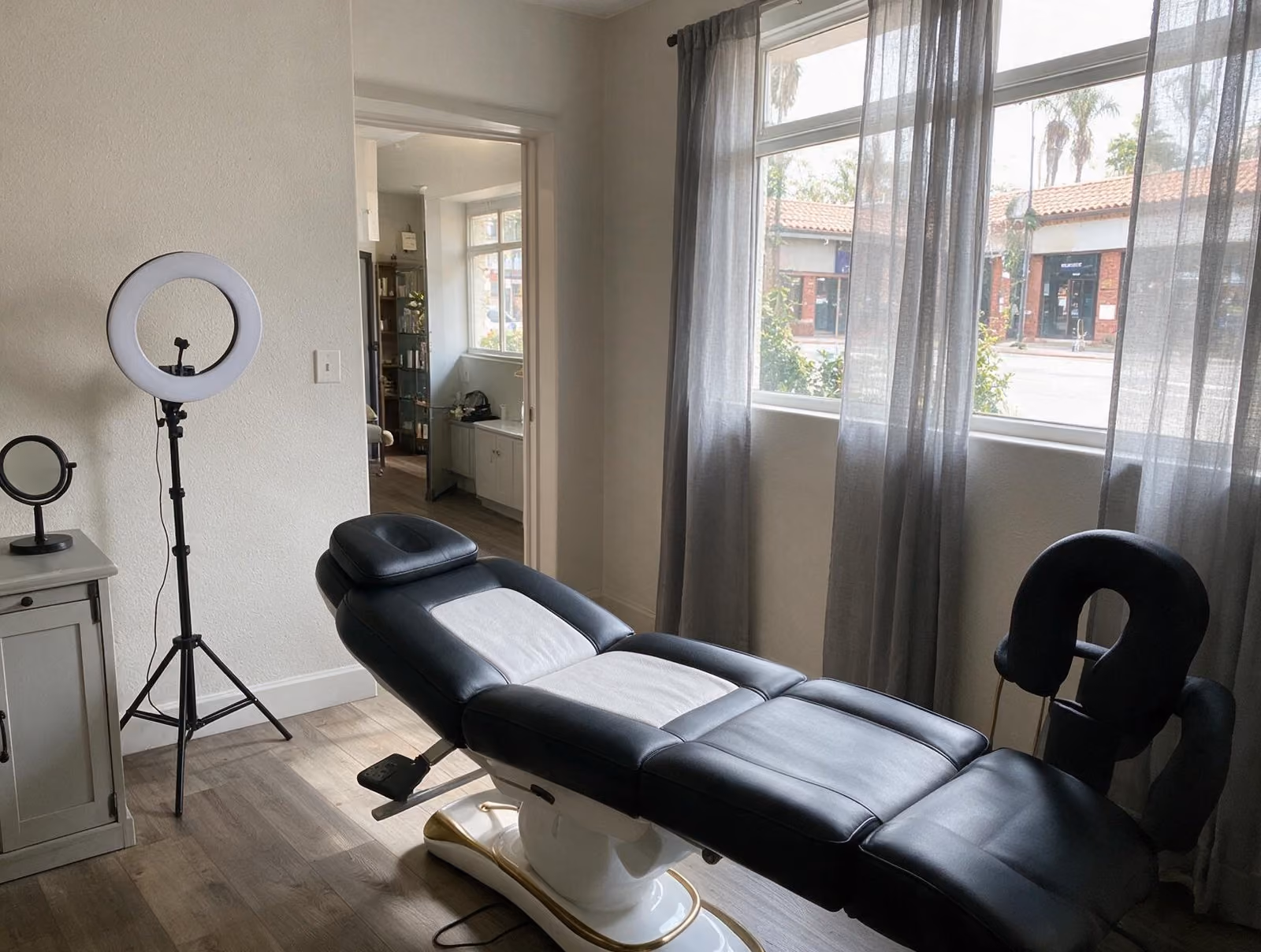 Treatment room with a black and white reclining treatment bed, a ring light on a tripod and sheer gray curtains