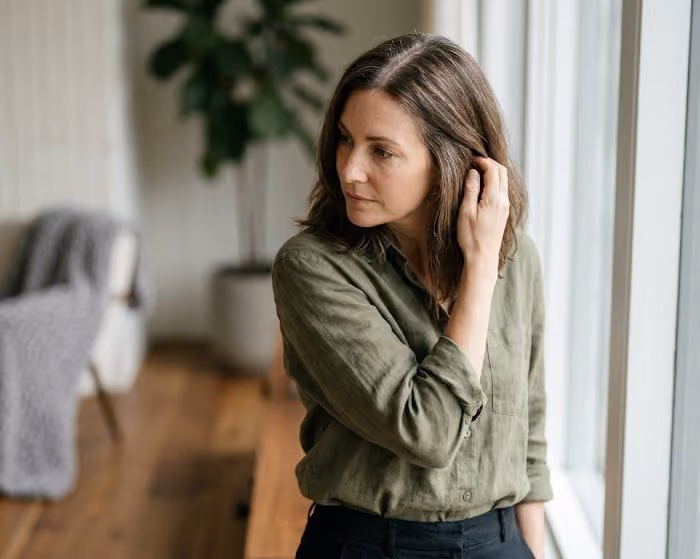 Woman with shoulder-length brown hair touching her hair as she looks out of a window in a bright room