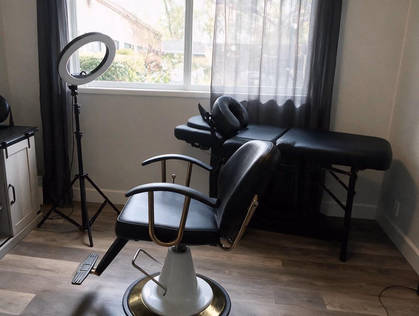 Treatment room with a black reclining chair, a ring light on a tripod and a black treatment bed by a window