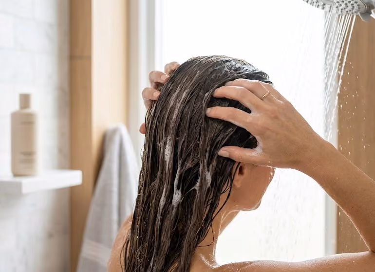 Woman with long wet brown hair washing her scalp with soapy hands under a shower