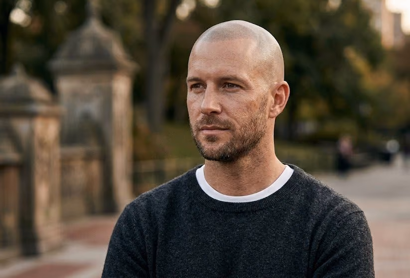 Man with a shaved head and short beard in a dark sweater, looking to the side in a park
