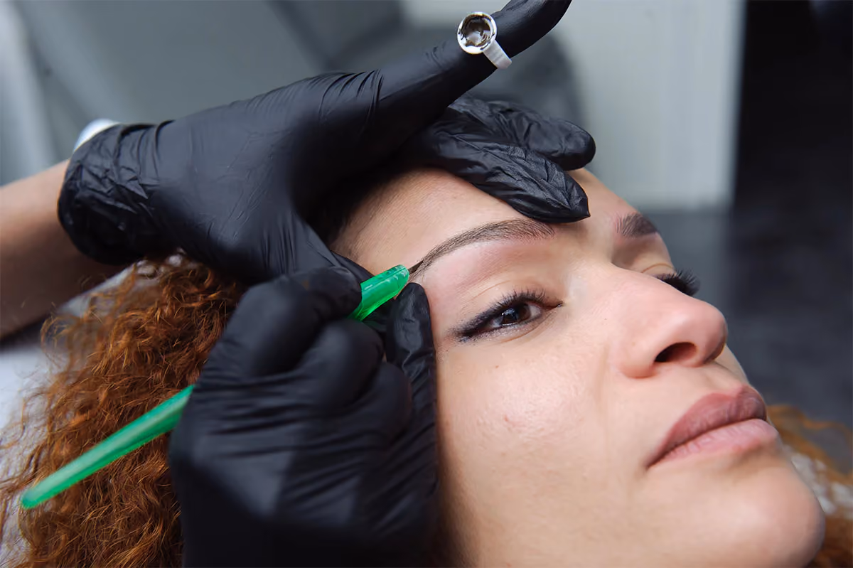 Gloved hands drawing a brow stroke with a green-handled tool on a woman with curly red hair