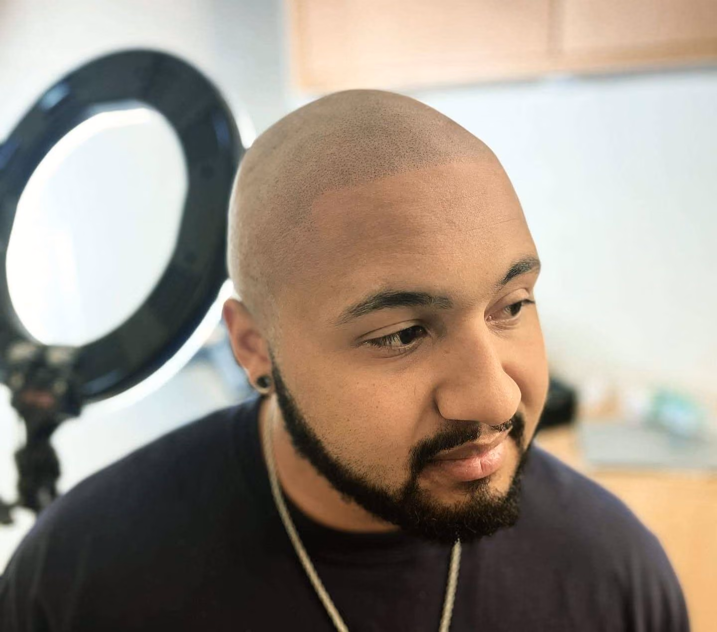 Man with a closely shaved head and light stubble looking to the side near a window, with a ring light behind him